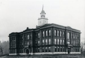Black and white image of a three story brick building with a clock tower in the middle, with a flag pole displaying the American flag on top.