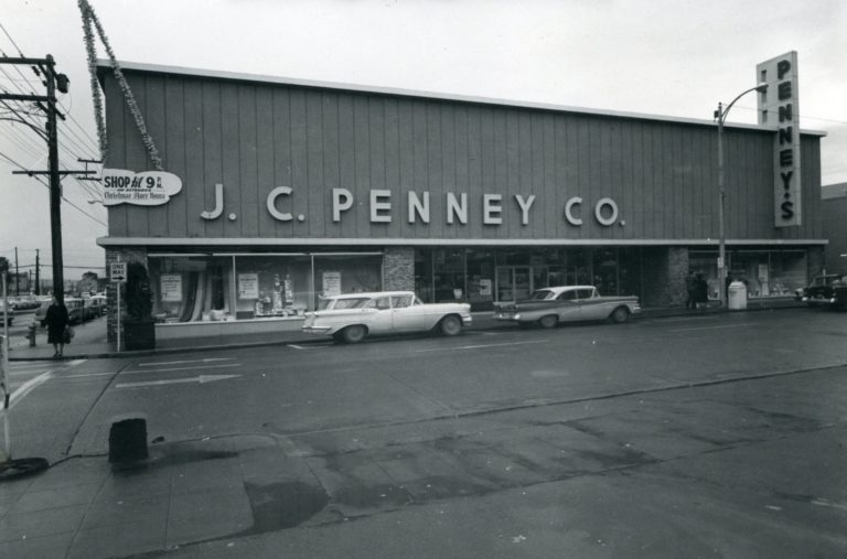 Black and white image of the J.C. Penney Co. two-story building, taken from across the street with two cars parked in front