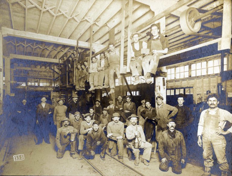 Black and white image of 37 men posed either kneeling, sitting, or standing inside of a large wooden building with rail tracks on the ground