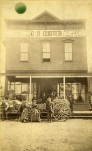 Sepia image of a two-story wooden building with a large sign reading, "C.S. Custer Groceries and Provisions, Dry Goods and Notions". Men and women are standing on the porch and in front of the building, as well as a horse-drawn carriage.