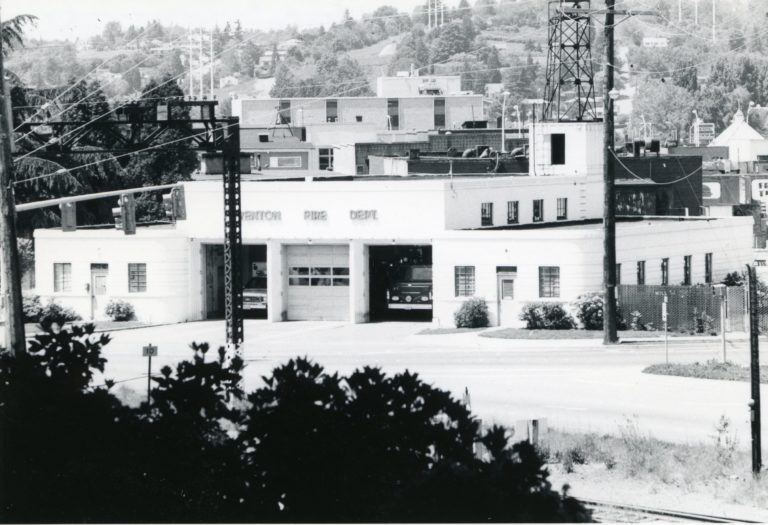Black and white image of an art-deco style building with one fire and one paramedic truck parked inside the first and second garage bays