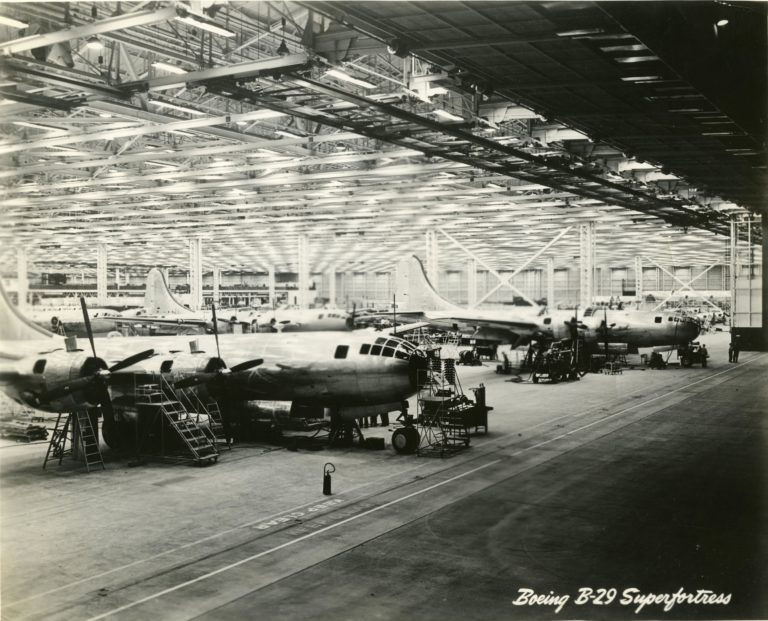 Black and white image of a large warehouse space. Four airplanes can be seen in various stage of completion.