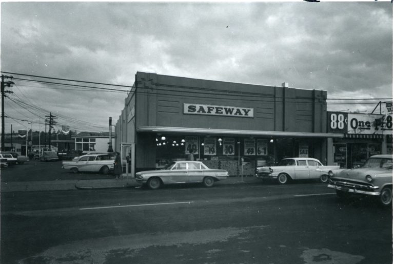 Black and white image of a two story building with an awing, a large sign the reads "Safeway", and price advertisements in the windows. Cars are parked on the street in front and in the parking lot to the left of the building.
