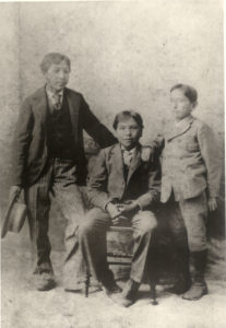 A black and white image of three boys of varying ages wearing suits. The boy in the middle is sitting in a chair and the one on the left is holding a hat.