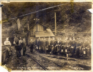 Black and white image of fifteen men sitting or standing outside a coal mine. Rail tracks are in the foreground and a donkey is nearby.