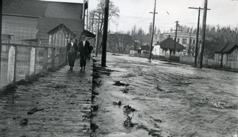 Black and white image of two boys walking down a wooden sidewalk with the town streets beside them flooded