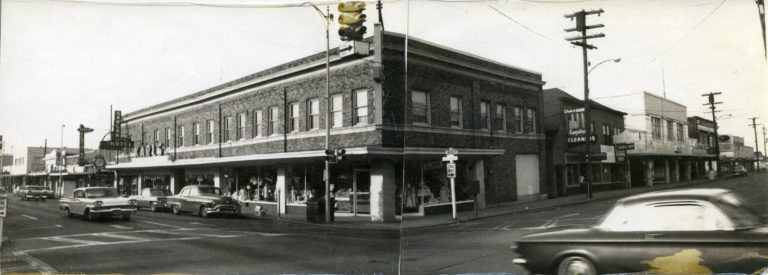 Black and white image of a two-story brick building with other commercial buildings on either side going down the block with cars driving on the street