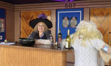 Woman behind a counter wearing a witch costume next to a scarecrow dressed in a wedding gown