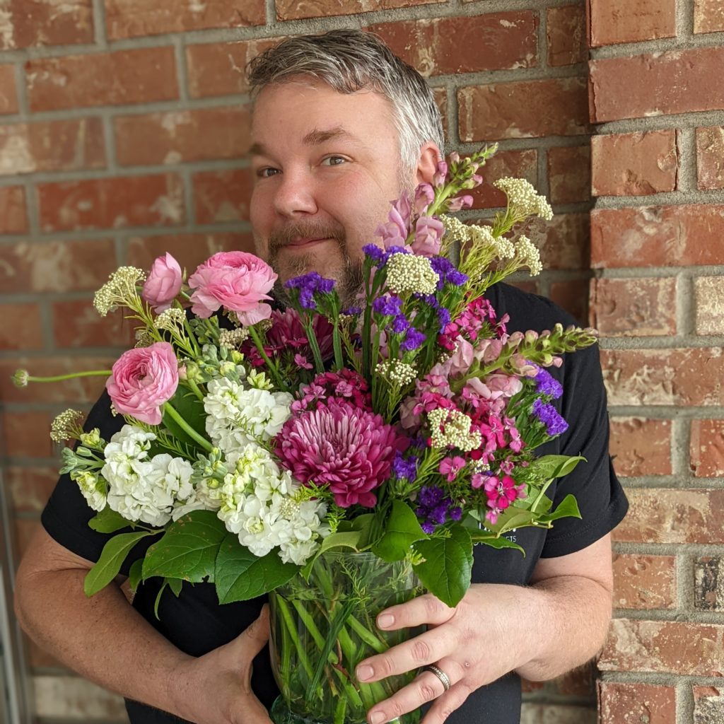 A man with a beard holds a large bouquet of flowers, there is a brick wall in the background