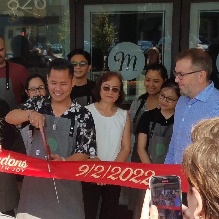 A group of people gathered in front of Macadon's shop entrance, with the owner cutting a red ribbon with oversized scissors.